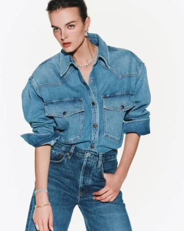 Model Aylah Peterson poses in a blue denim shirt and matching jeans paired with diamond jewelry against a white background.