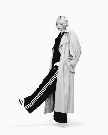 A woman with blonde hair poses in a long beige coat and black Adidas track pants with sneakers against a bright white backdrop.