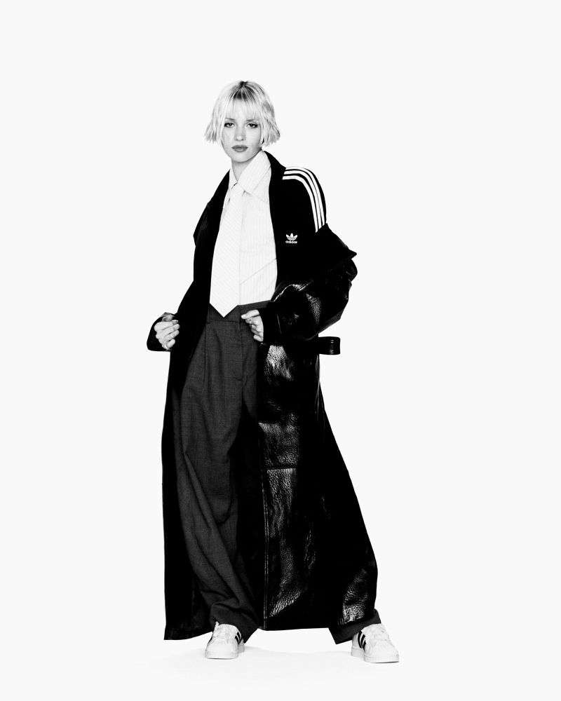 Full-length shot of a woman with blonde hair wearing a black leather coat, white shirt, grey pants, and white sneakers against a white background.