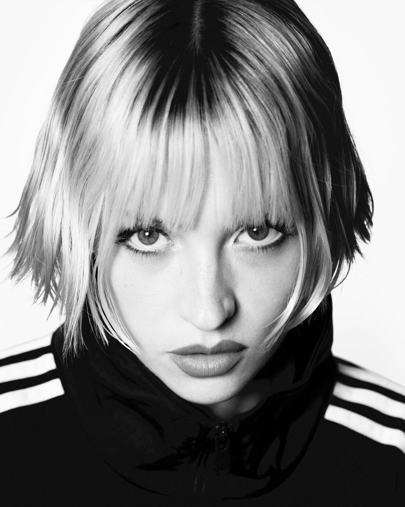 Close-up black and white portrait of Angle wearing an Adidas track jacket against a plain background.