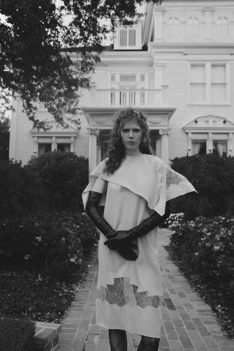 Monochrome portrait of a woman in a dress with lace details, wearing long gloves, posing outside a large house with manicured landscaping.