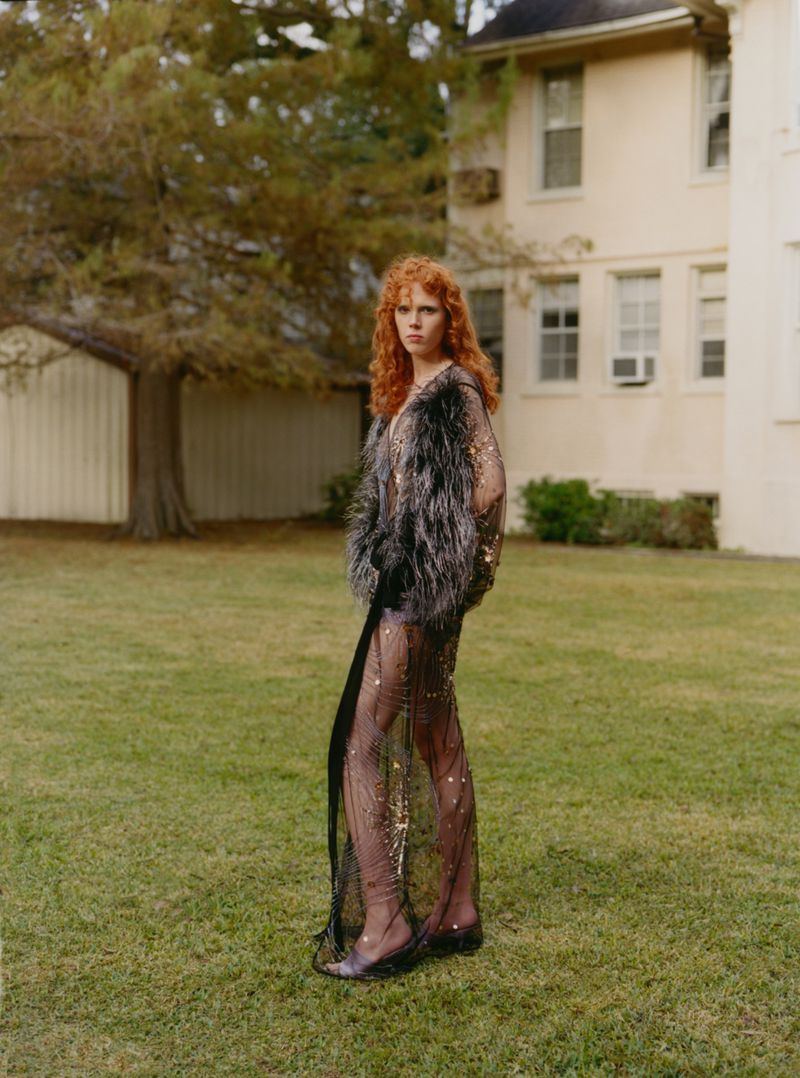 Red-haired woman in semi-sheer sequined dress and fur jacket poses on a lawn in front of a two-story beige building on a cloudy day.