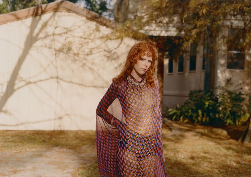 Adult female with red hair poses in front of a beige building, wearing a patterned sheer dress outdoors in a fashion editorial style.