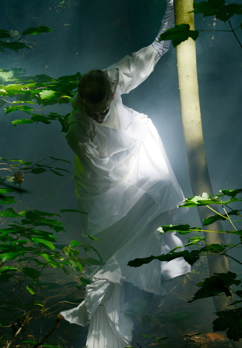 Woman in sheer white costume embracing tree in misty forest. Low-angle, full shot. Sunlight breaks through canopy creating ethereal effect.