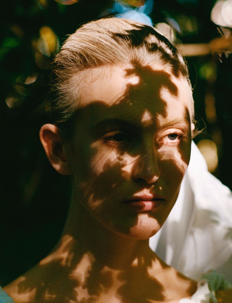Close-up portrait of a fair-skinned young woman with light blonde hair, with her face partially shaded by leaf shadows in a natural outdoor setting.