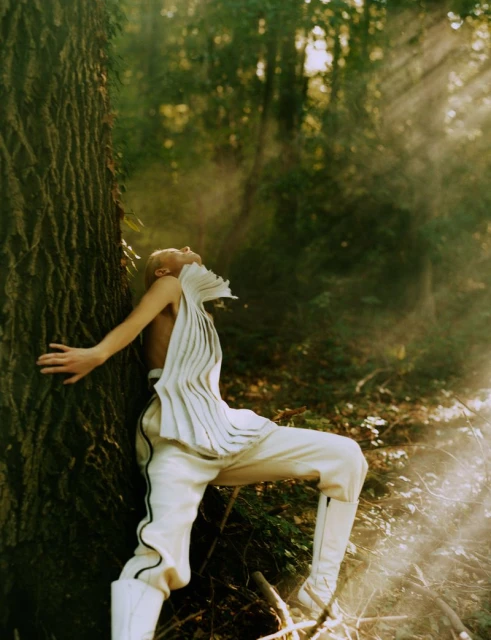 Woman in stylized white clothing leans against a tree in sun-dappled forest. Fashion editorial in nature.