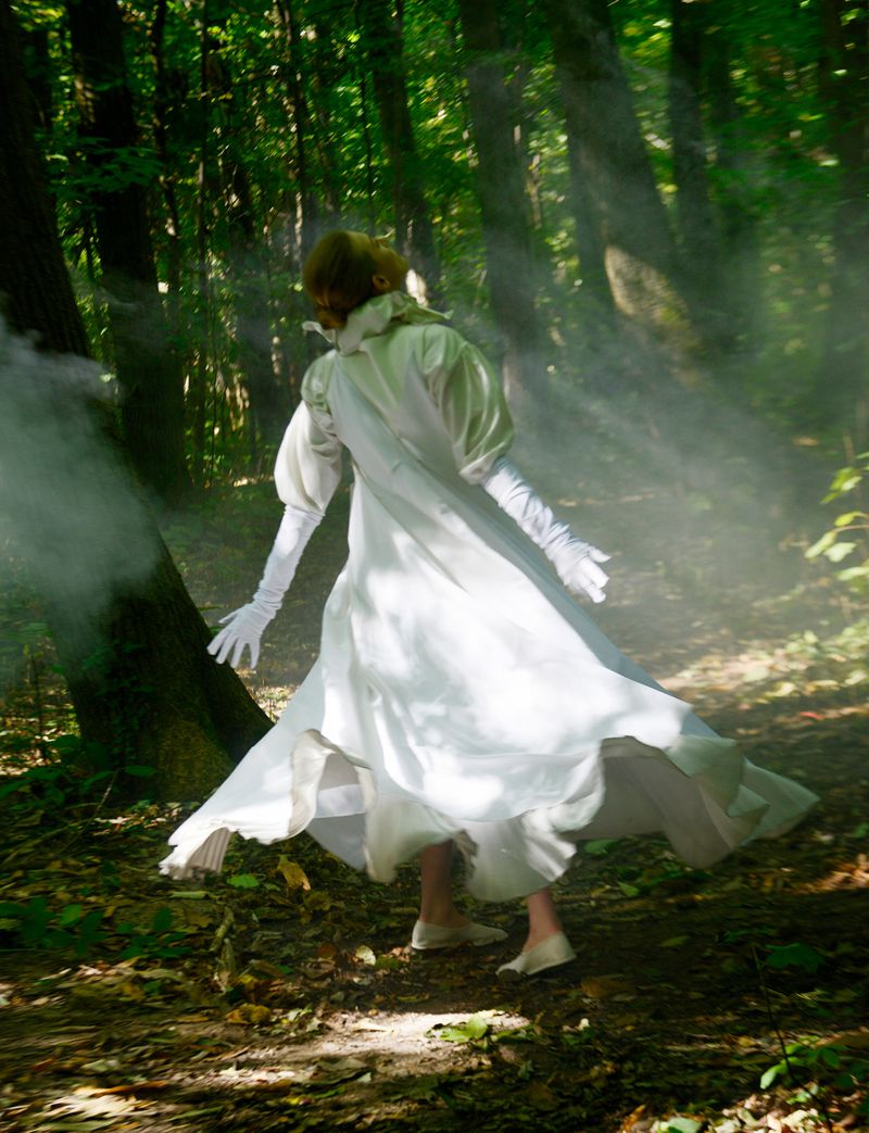 A woman in a white dress stands in a sunlit forest with a mist or smoke effect, creating an ethereal and slightly mysterious scene.