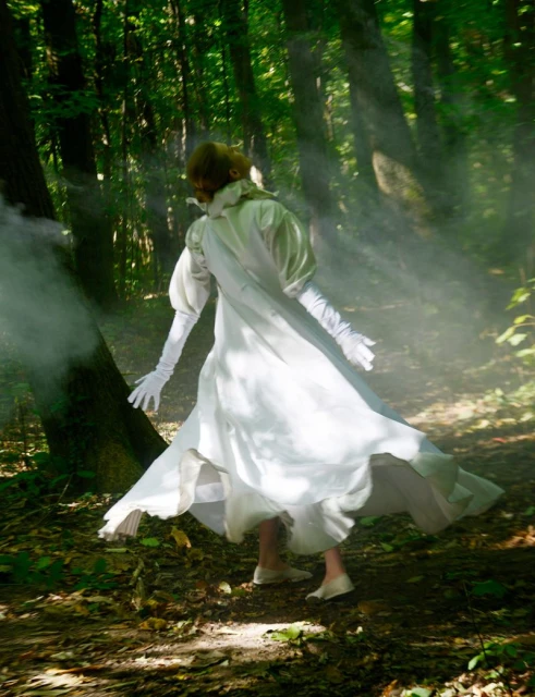 A woman in a white dress stands in a sunlit forest with a mist or smoke effect, creating an ethereal and slightly mysterious scene.