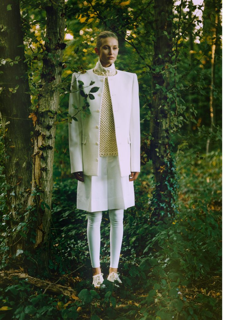 A woman in a white blazer and skirt poses in a lush, green forest setting. The woman stands, surrounded by trees and foliage.