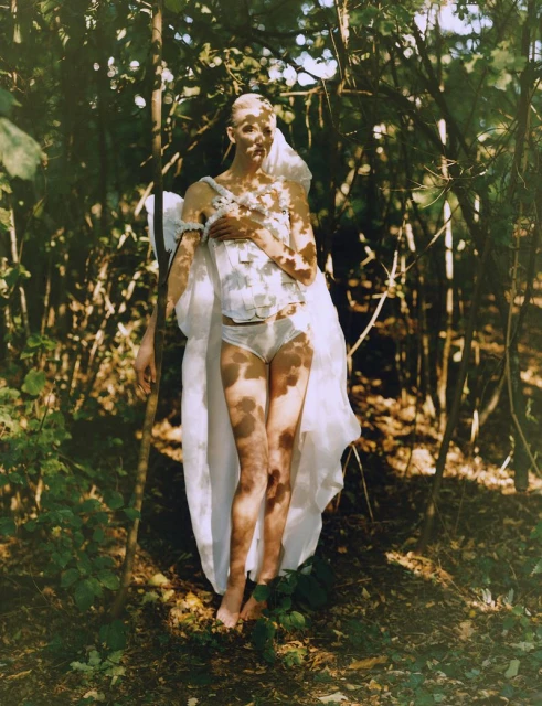 Fashion shot of a bare-footed young adult female model in white lingerie and a long, white gown standing outdoors in a wooded area.