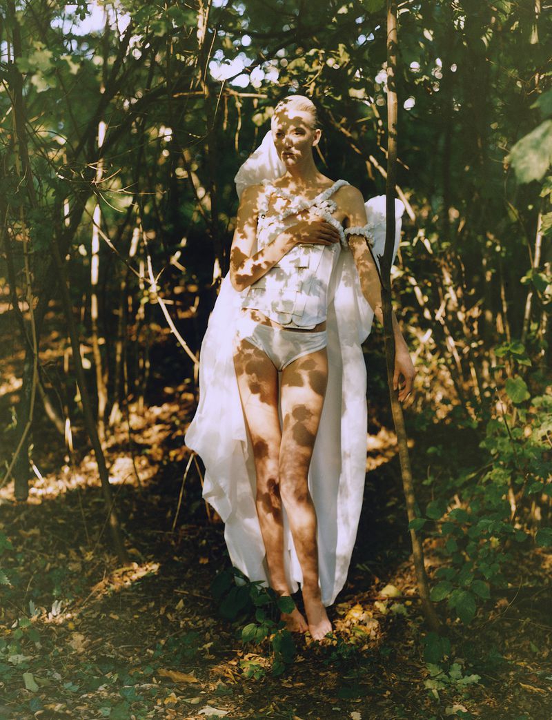 Woman in white corset and bottoms poses in a sun-dappled forest, surrounded by green foliage and slender tree trunks in artistic image.