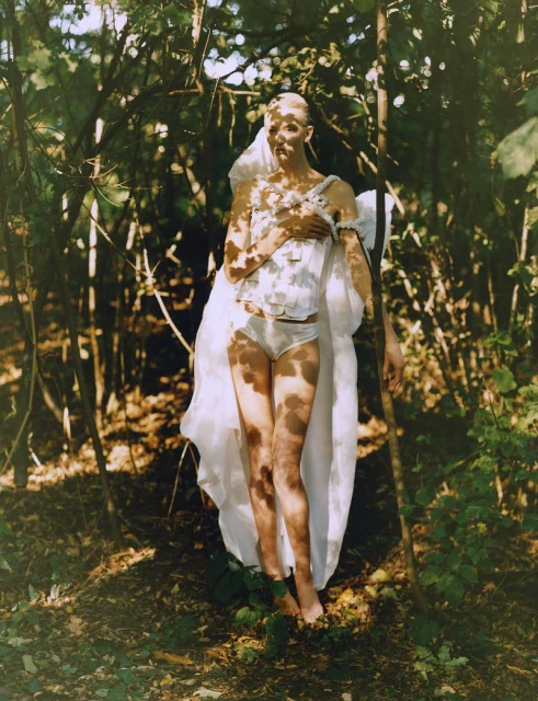Woman in white corset and bottoms poses in a sun-dappled forest, surrounded by green foliage and slender tree trunks in artistic image.