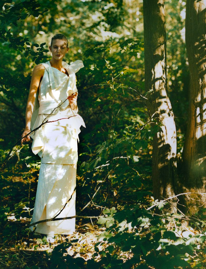 A woman in a cream-colored dress stands in a forest, surrounded by lush greenery and dappled sunlight filtering through the trees.