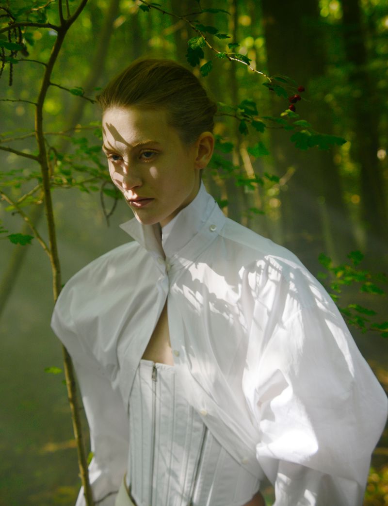 Woman in white standing in forest. Fashion shot with green foliage and soft lighting. Corset and puffy shirt. Mysterious mood.