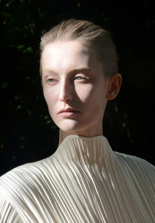 Model Alayna Neaum wearing a cream pleated high-neck top in directional sunlight against a dark foliage background.