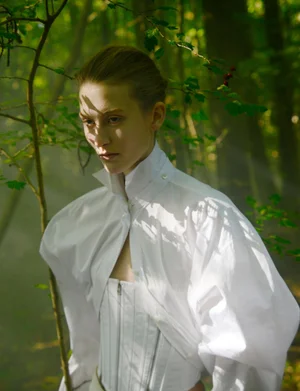 Alayna Neaum wears an architectural white blouse and corset with dappled shadows in a green woodland setting.