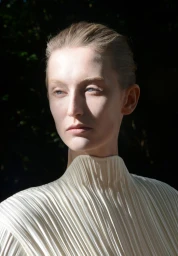 Model Alayna Neaum wearing a cream pleated high-neck top in directional sunlight against a dark foliage background.