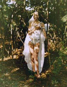 Model Alayna Neaum wearing white draped corsetry and briefs, standing barefoot among trees with dappled sunlight shadows.