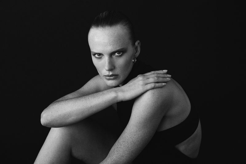 Monochrome portrait of a fair-skinned woman with slicked-back hair, seated with arms crossed, gazing directly at the camera against a black background.