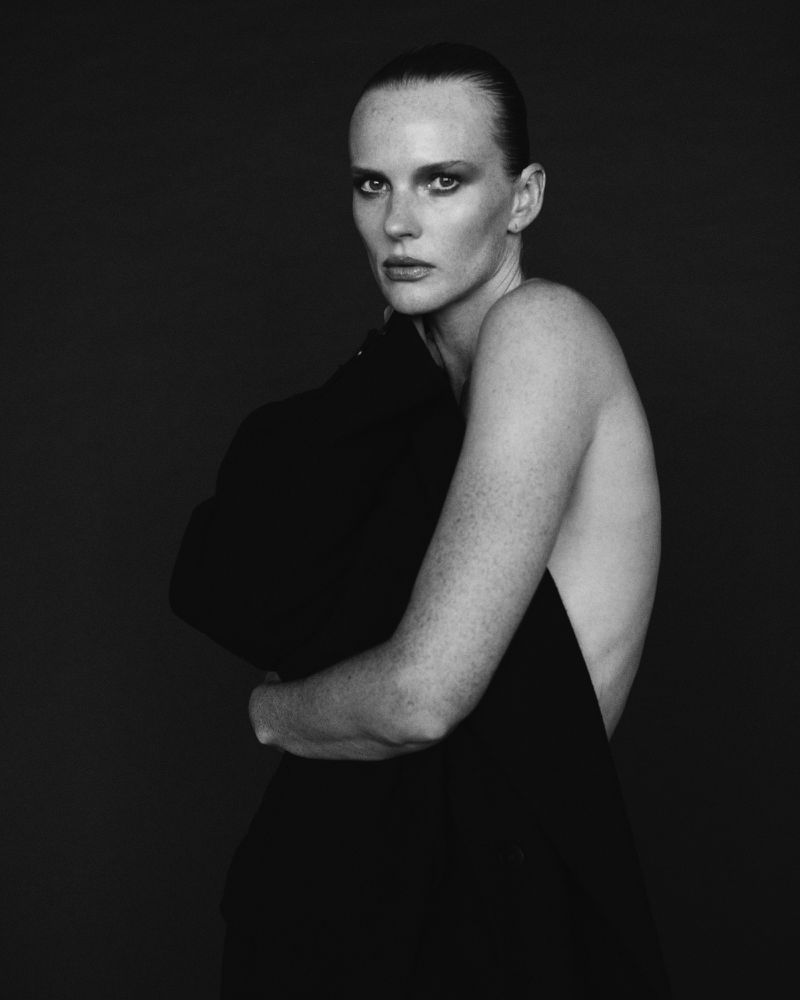 Black and white portrait of a fair-skinned, freckled woman with dark hair pulled back, draped in a dark blazer against a dark background.
