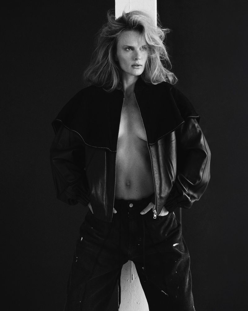 Black and white studio portrait of a blonde woman in a leather jacket and jeans with her hands on her hips in front of a pillar.