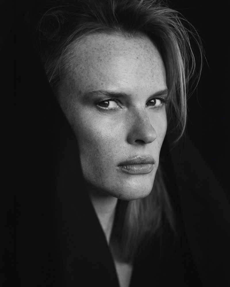 Black and white close-up portrait of model Anne Vyalitsyna framed by a dark hood looking directly forward.