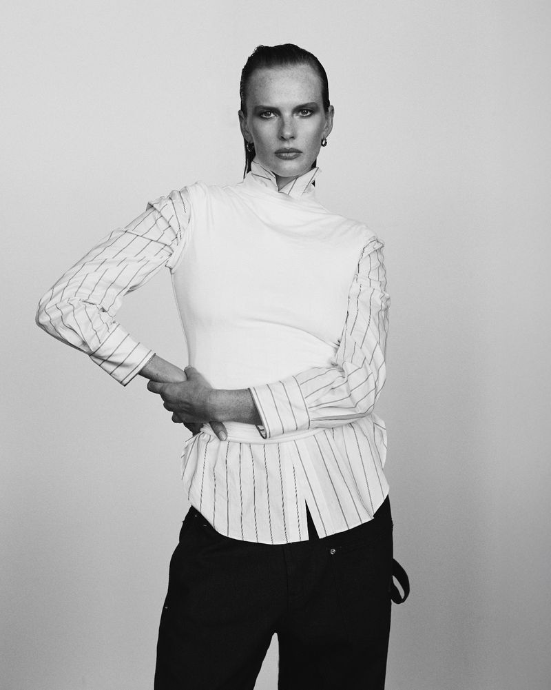 Black and white portrait of a fashion model with slicked-back hair wearing a white top layered over a pinstripe shirt and dark pants against a gray backdrop.