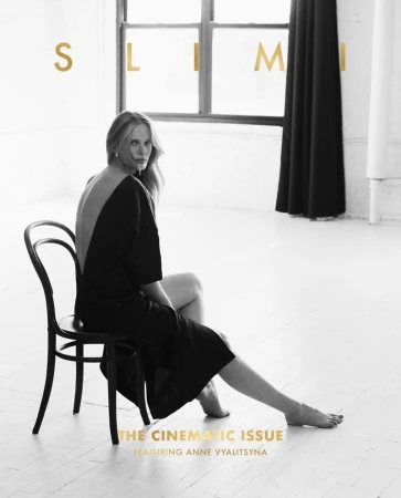 Elegant fashion shot featuring a poised blonde model in a black dress seated on a chair in a minimalist white photo studio setting.