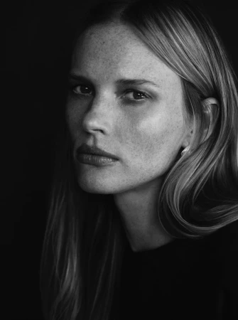 Black and white close-up portrait of model Anne Vyalitsyna with freckled skin and dark clothing against a black background.