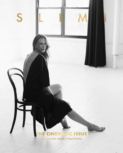 Model Anne Vyalitsyna seated on a dark wooden chair in a black dress inside a bright studio interior for SLIMI Magazine.