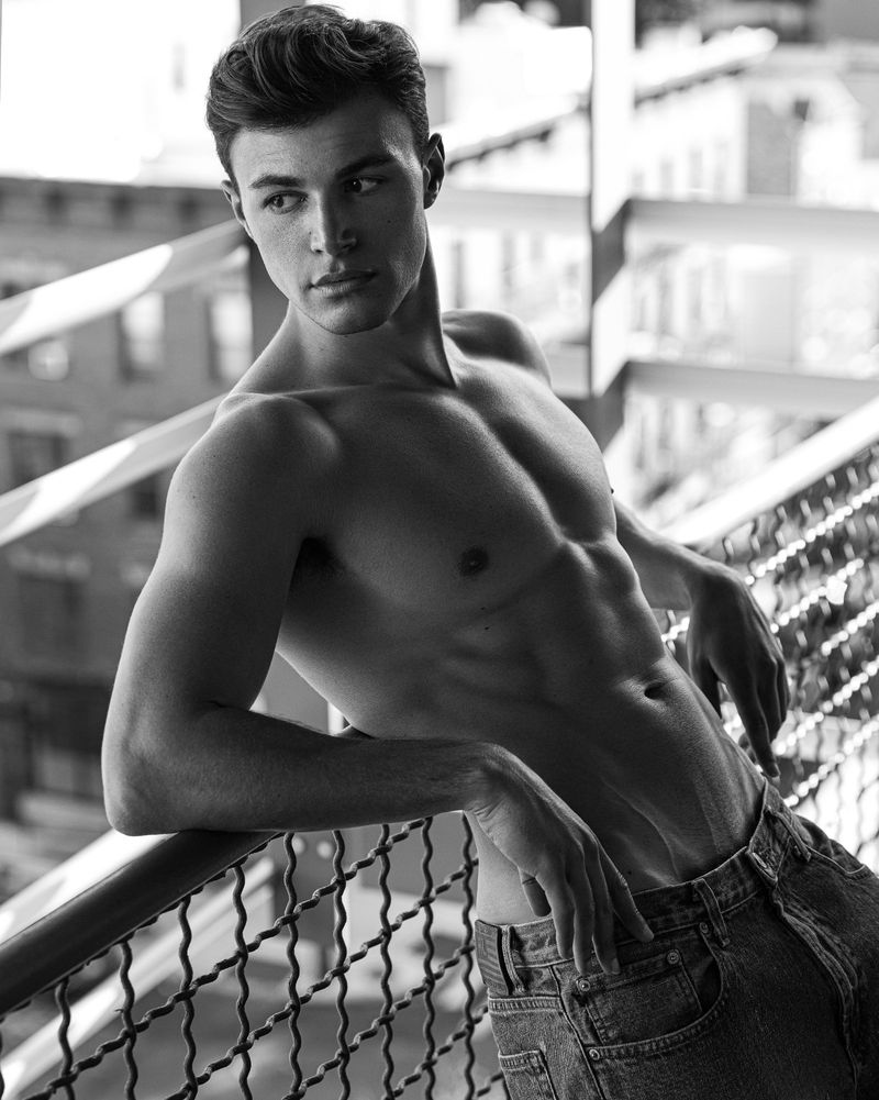 Black and white portrait of a shirtless adult male with dark hair, wearing jeans, leaning on a railing, with an urban background.