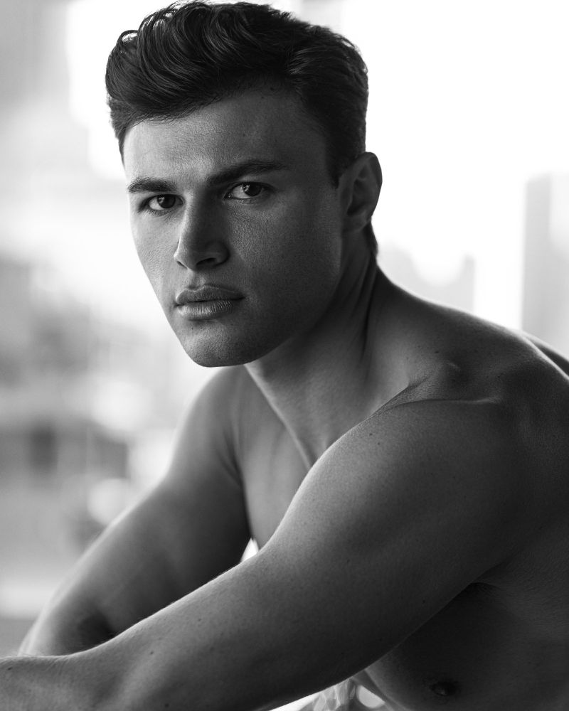 Black and white portrait of a shirtless man with styled dark hair looking seriously at the camera.