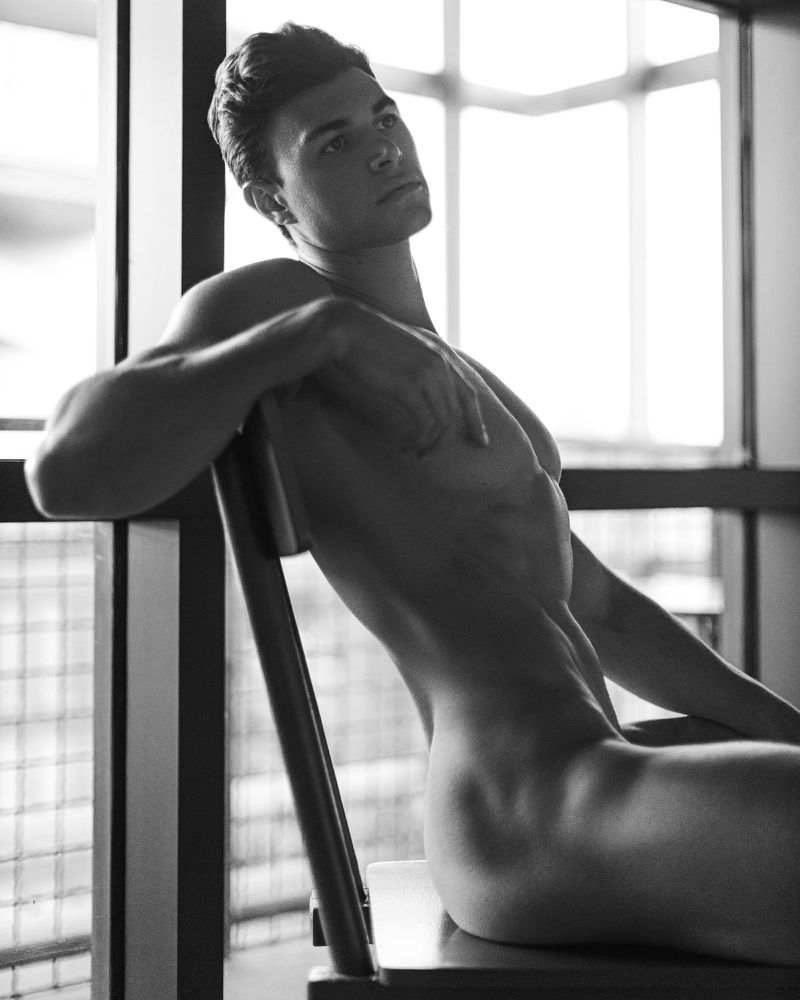 Monochrome portrait of shirtless man sitting on wooden chair near window with natural light, showcasing muscular physique and stylish hair.