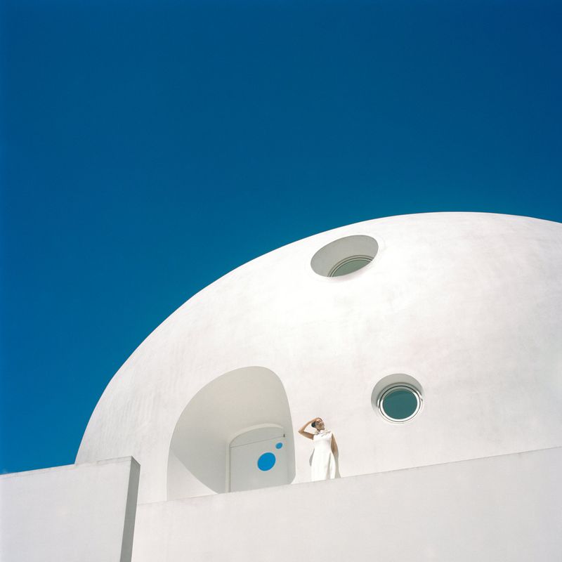 Woman in white dress stands near modern, white, domed building against a clear blue sky on a bright sunny day.