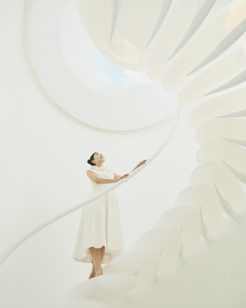 Adult woman in white dress on all-white spiral staircase looking upwards, bathed in bright and soft light, creating an ethereal atmosphere.