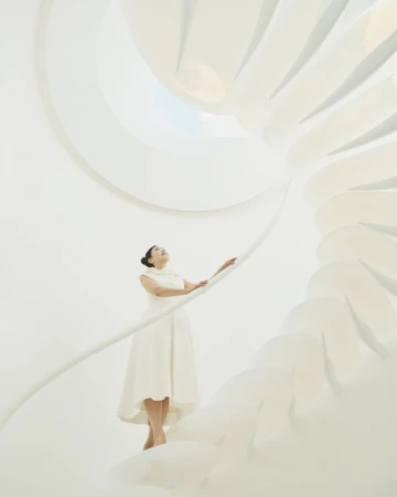 Adult woman in white dress on all-white spiral staircase looking upwards, bathed in bright and soft light, creating an ethereal atmosphere.