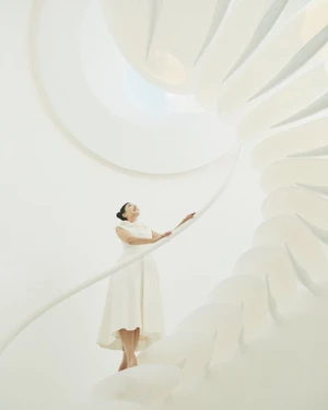 East Asian woman in an off-white midi dress standing on a sculptural white spiral staircase looking up toward a light source.