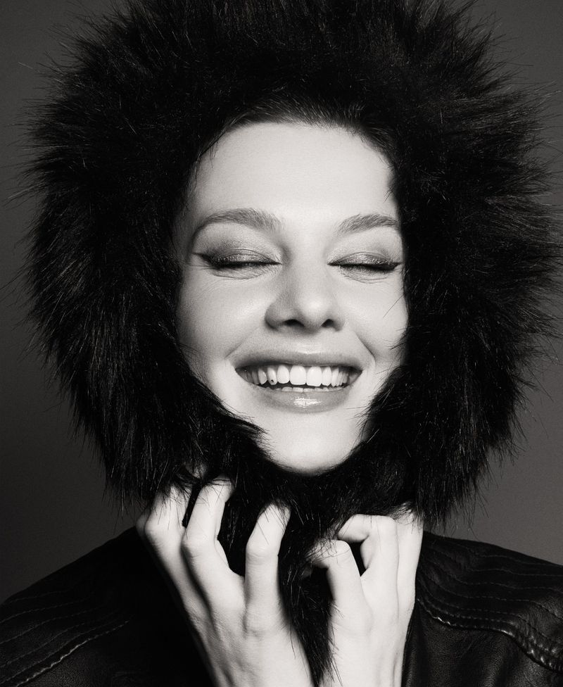 Monochrome portrait of a smiling adult woman in a fur-lined jacket, her hands playfully touching the fur around her face, eyes closed.