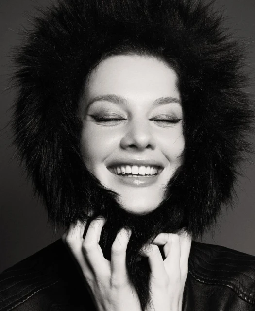 Monochrome portrait of a smiling adult woman in a fur-lined jacket, her hands playfully touching the fur around her face, eyes closed.