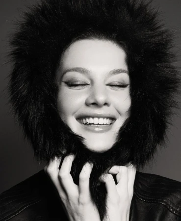 Monochrome portrait of a smiling adult woman in a fur-lined jacket, her hands playfully touching the fur around her face, eyes closed.