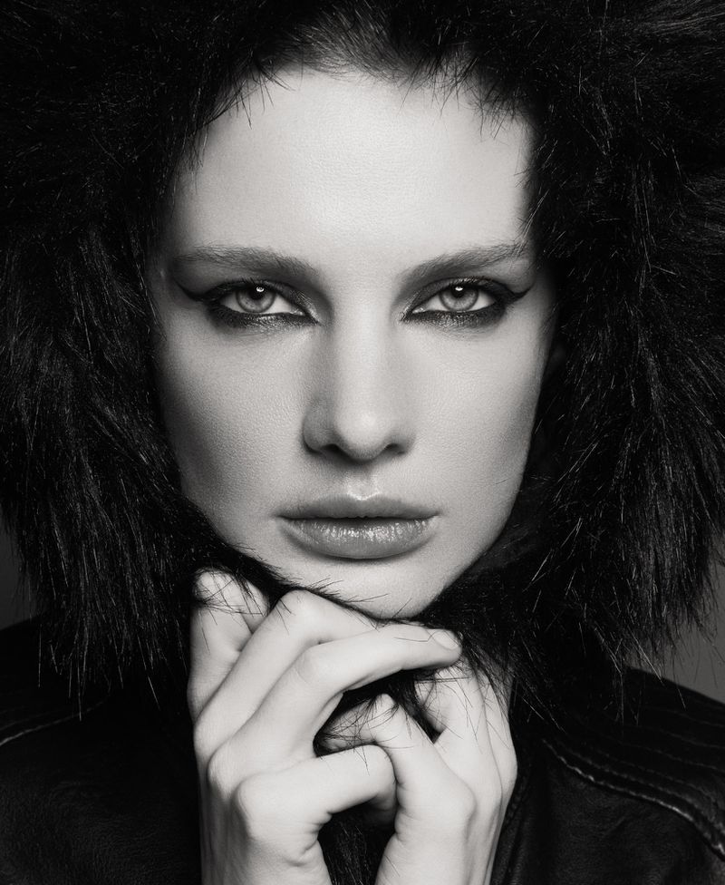 Striking black and white portrait of a woman with dark hair, dramatic eye makeup, and black leather jacket. Hands are seen near her neck.