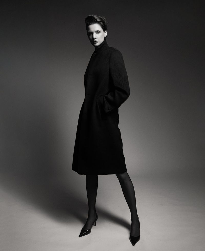 Elegant woman in black coat poses in a studio setting, showcasing fashion and style with neutral background, creating a sophisticated portrait.