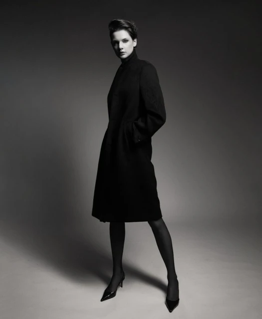 Elegant woman in black coat poses in a studio setting, showcasing fashion and style with neutral background, creating a sophisticated portrait.
