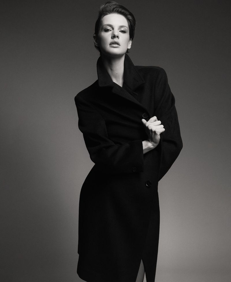 Monochrome portrait of a brunette woman in a black wool coat posing in front of a grey backdrop in a studio with soft lighting.
