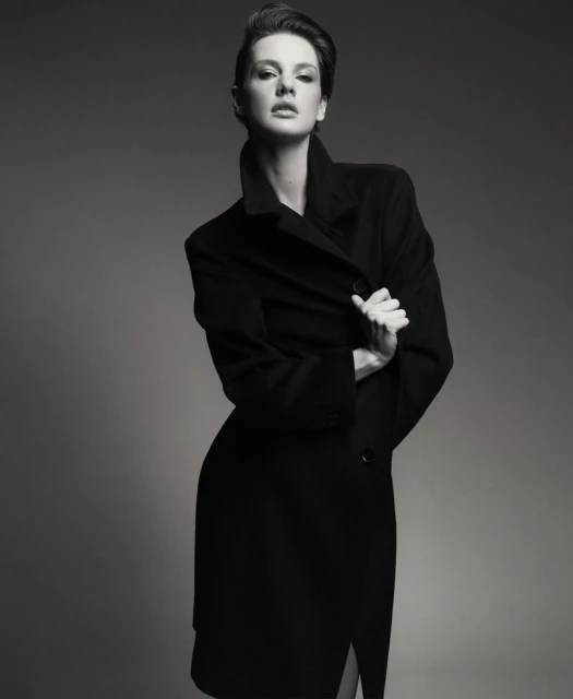 Monochrome portrait of a brunette woman in a black wool coat posing in front of a grey backdrop in a studio with soft lighting.