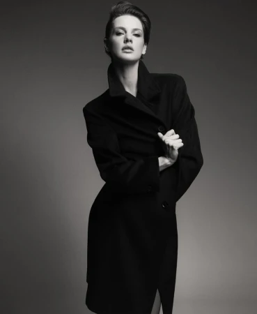 Monochrome portrait of a brunette woman in a black wool coat posing in front of a grey backdrop in a studio with soft lighting.