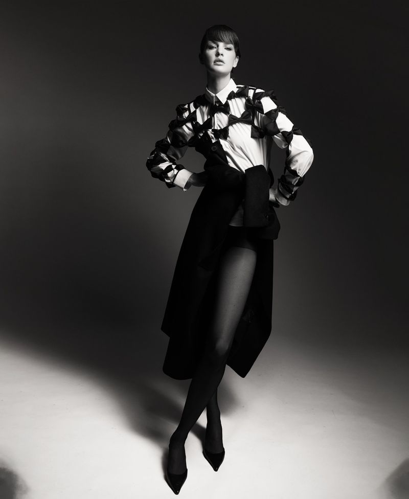 Model Mirthe Dijkstra wearing a bow-embellished collared shirt, black skirt, tights, and heels in a black and white studio portrait.