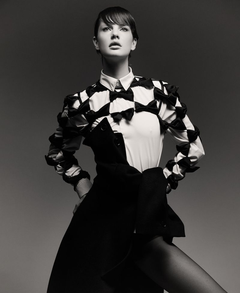 Black-and-white portrait of a woman in a collared shirt with bow details, and a dark jacket draped, against a grey backdrop.