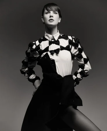 Black-and-white portrait of a woman in a collared shirt with bow details, and a dark jacket draped, against a grey backdrop.