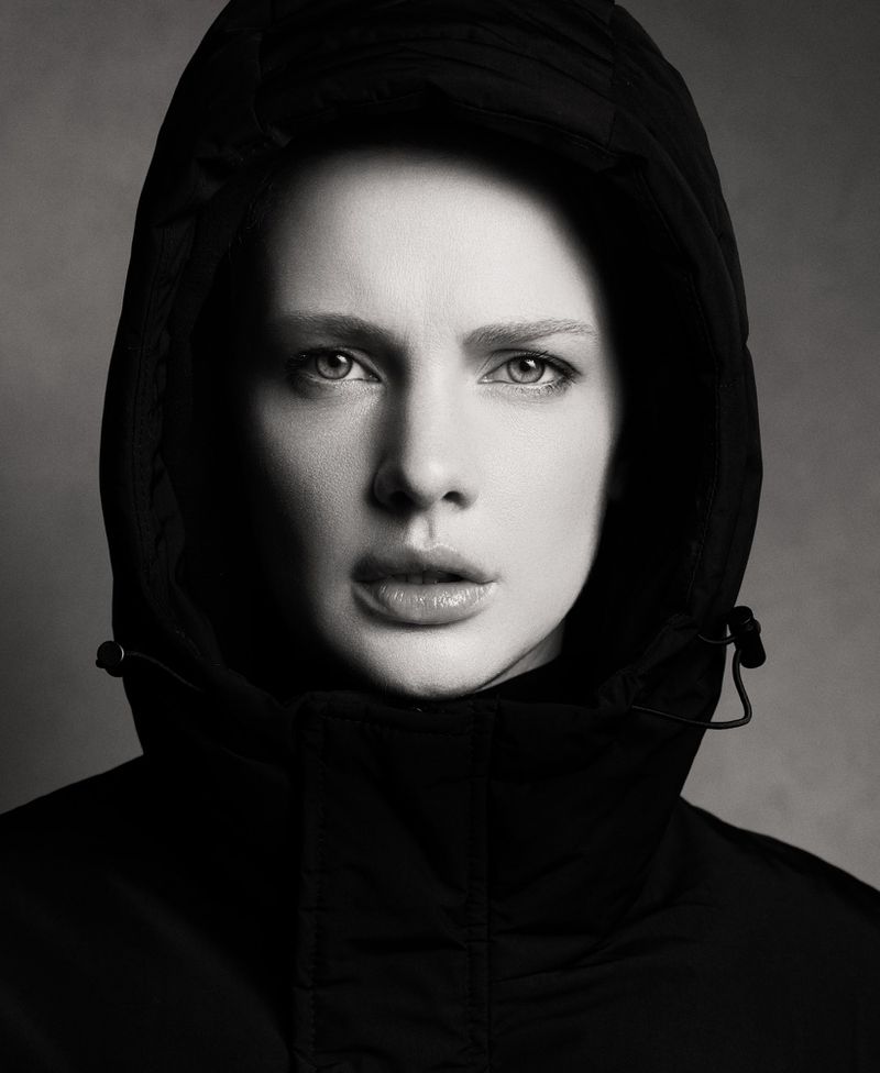 Monochrome portrait of a fair-skinned adult female with a hooded coat, face forward, with neutral expression against a grey background.